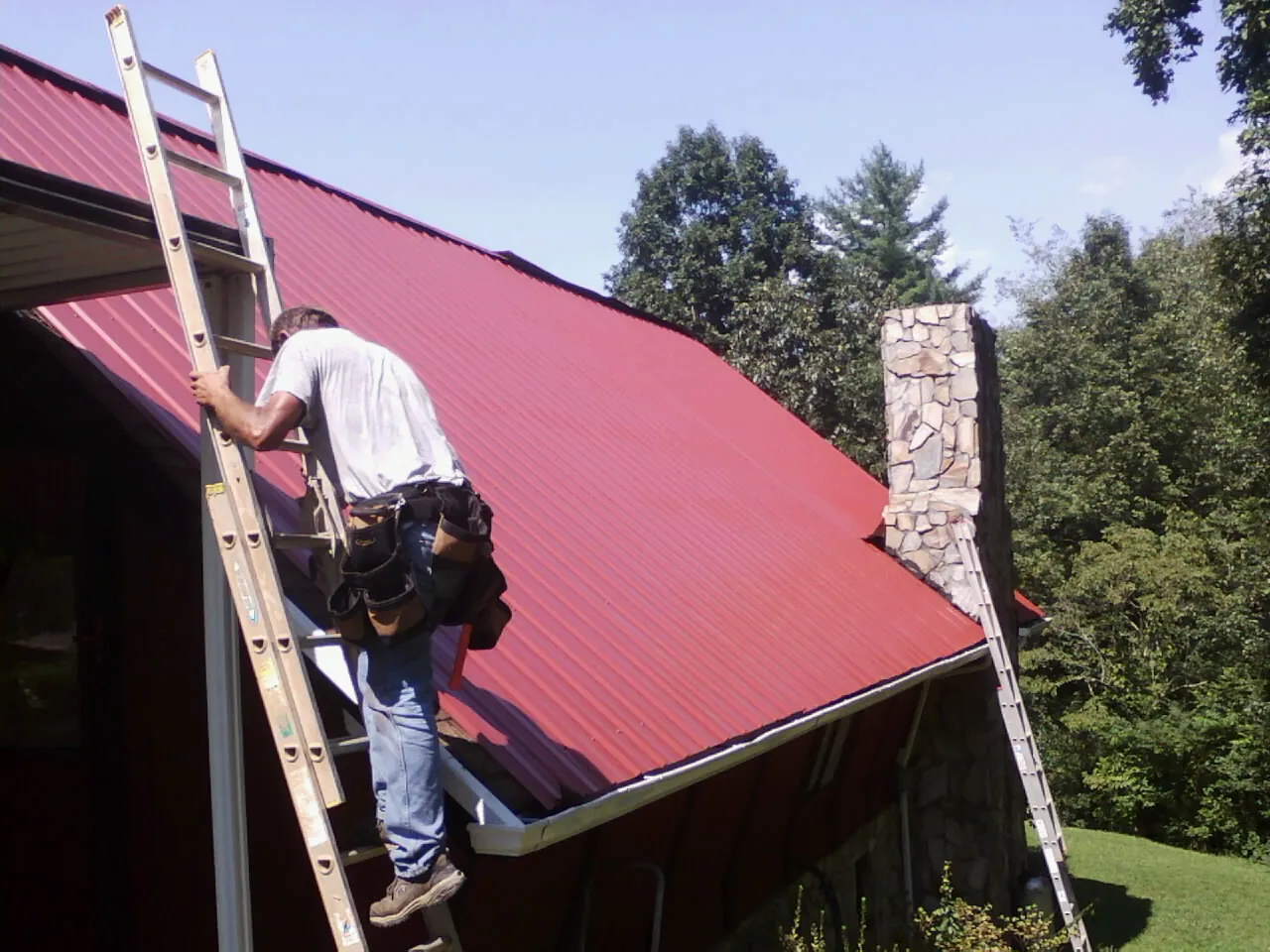 Roofer on a ladder beside a red metal roof and stone chimney — Soffit & Fascia Repair by Roofing Brothers in Spring Green